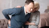 Man holds his back in his pain, as woman helps him stand up